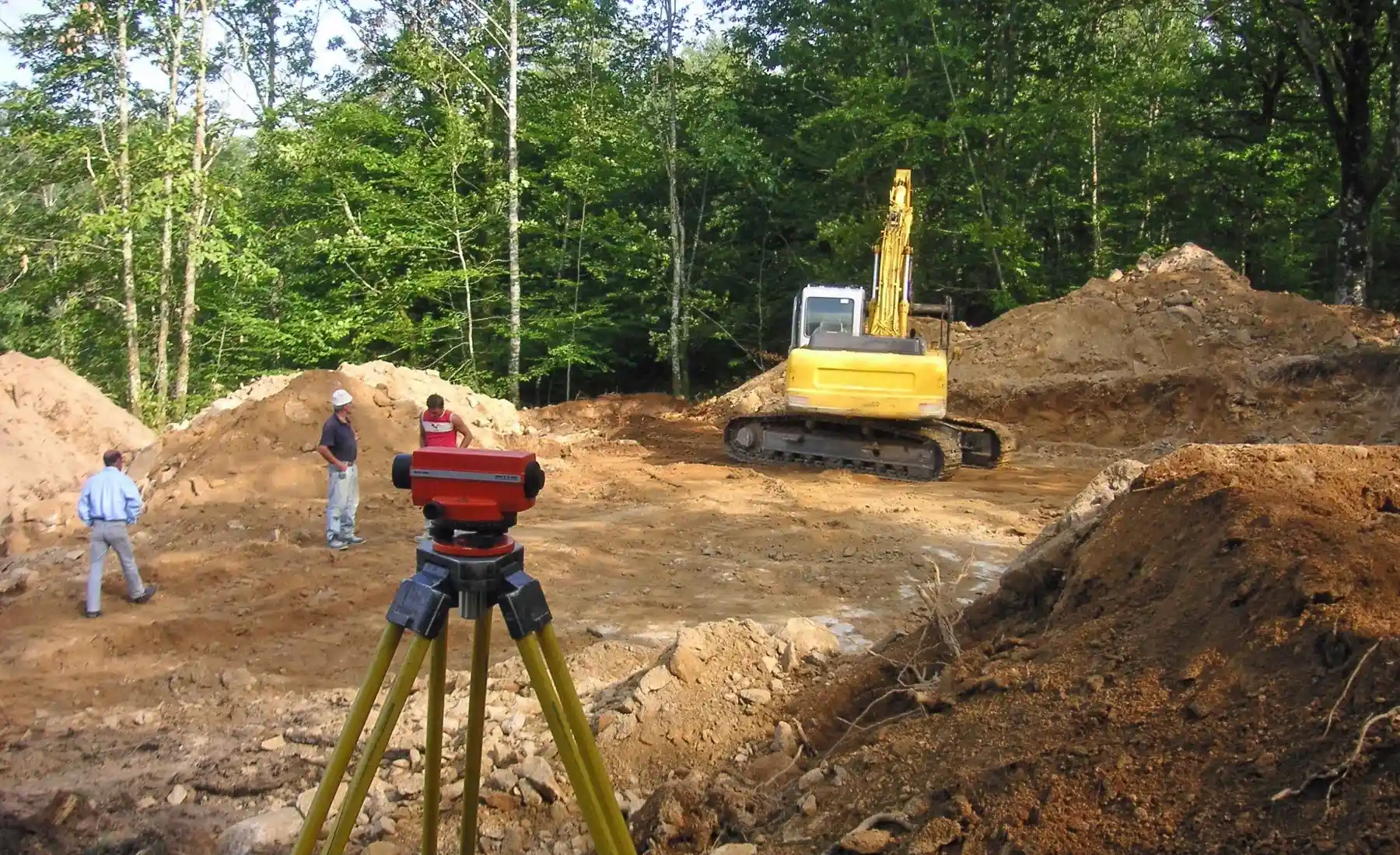 Construction site with surveying equipment in the foreground, workers discussing plans, and a yellow excavator operating near piles of soil surrounded by trees.